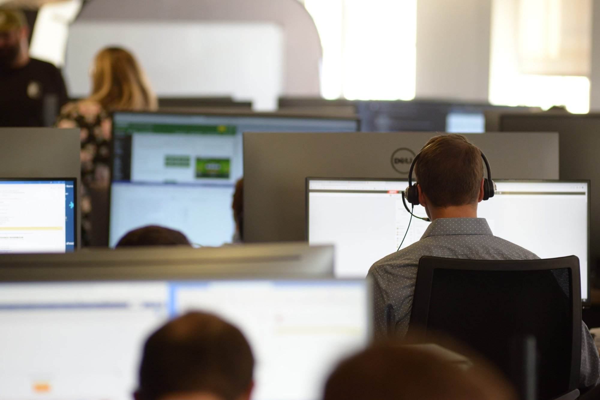 A man wearing a headset works at a desk in a busy office, surrounded by multiple computer screens and colleagues in the background.