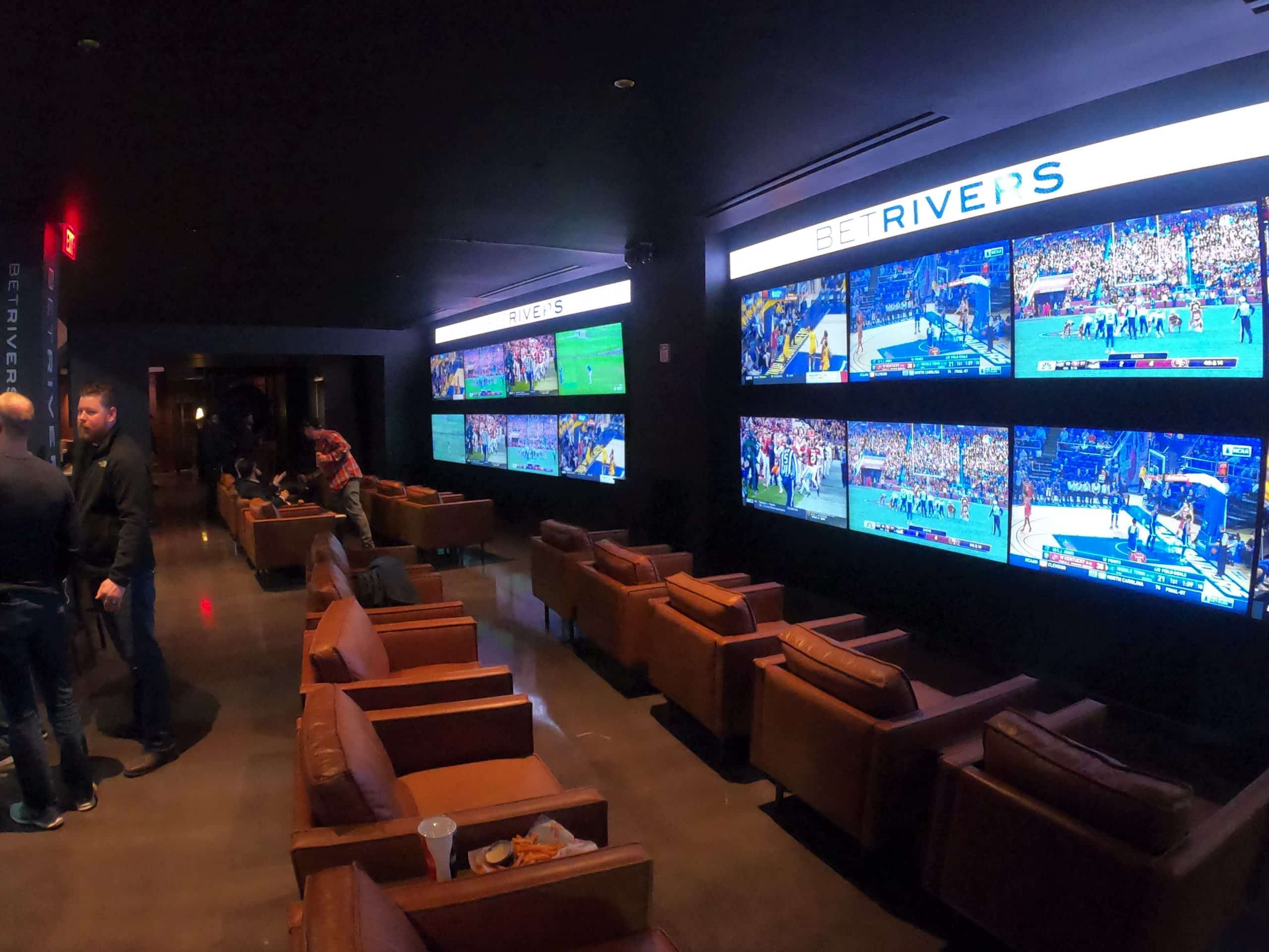 A spacious room filled with multiple televisions and comfy chairs with groups of people engaged in conversation around them.