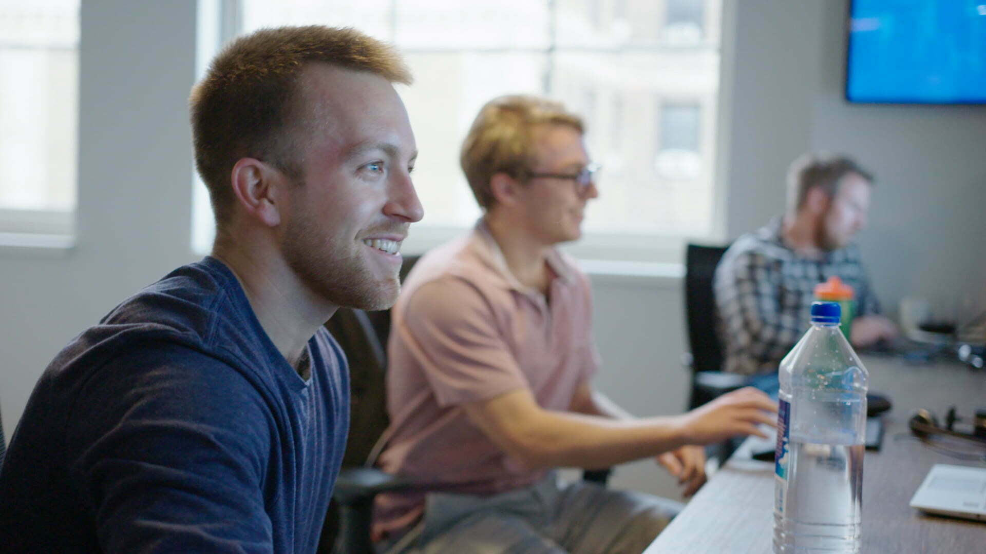 A man seated at a table with two other men, engaged in conversation and looking at laptops.