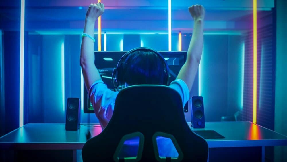 A gamer celebrates victory, raising arms in triumph while seated at a desk with a colourful, neon-lit background and gaming setup.