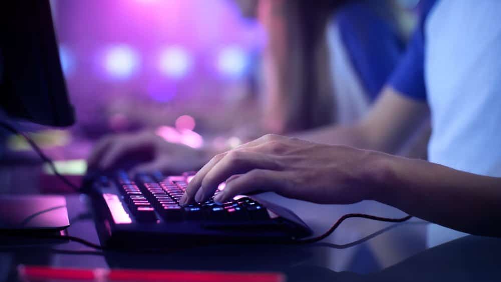 A close-up of a person's hand typing on a backlit keyboard in a dimly lit gaming environment with colourful lighting.