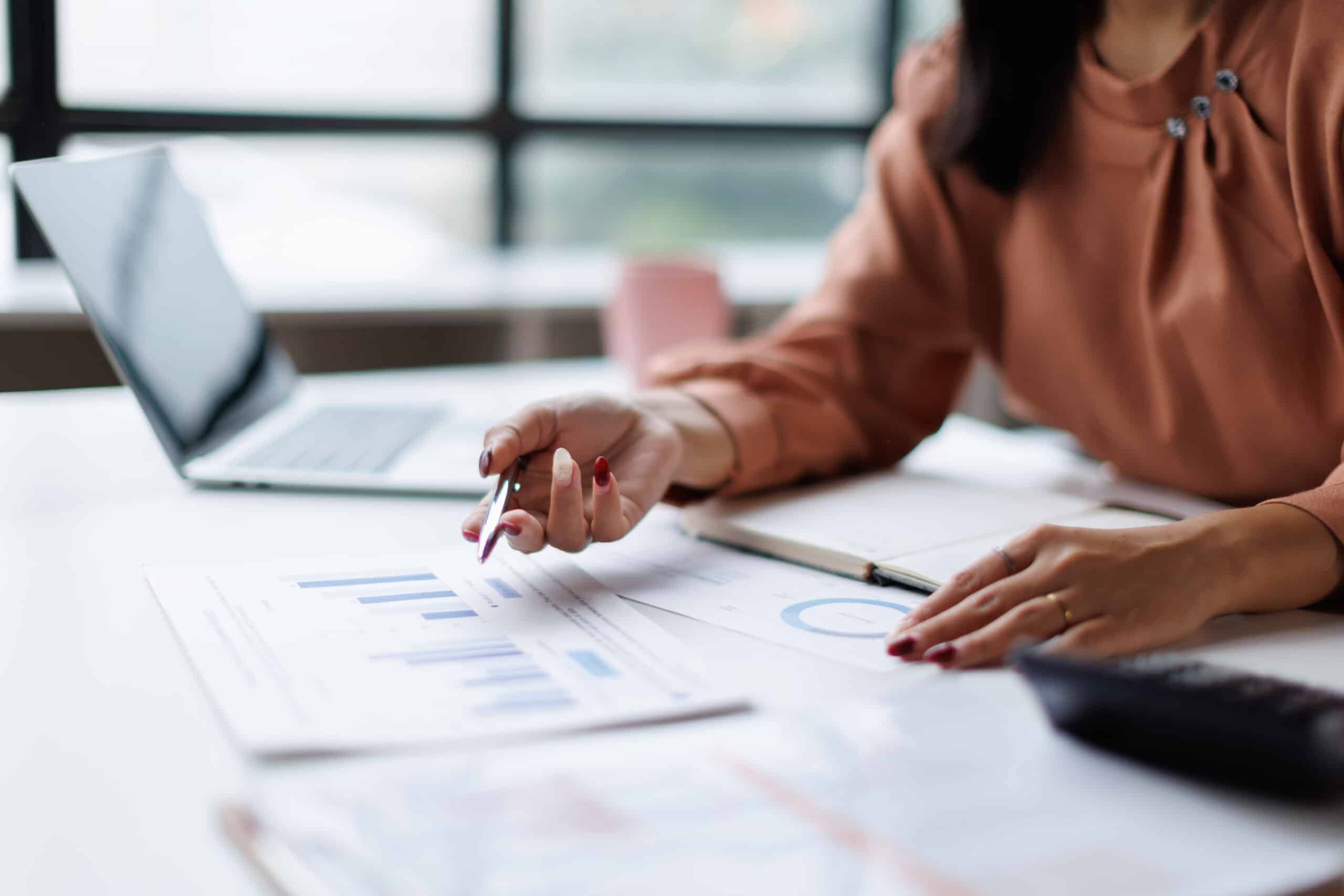Professional woman analyzing financial data and business reports, using a pen to point at charts while working on a laptop at an office desk